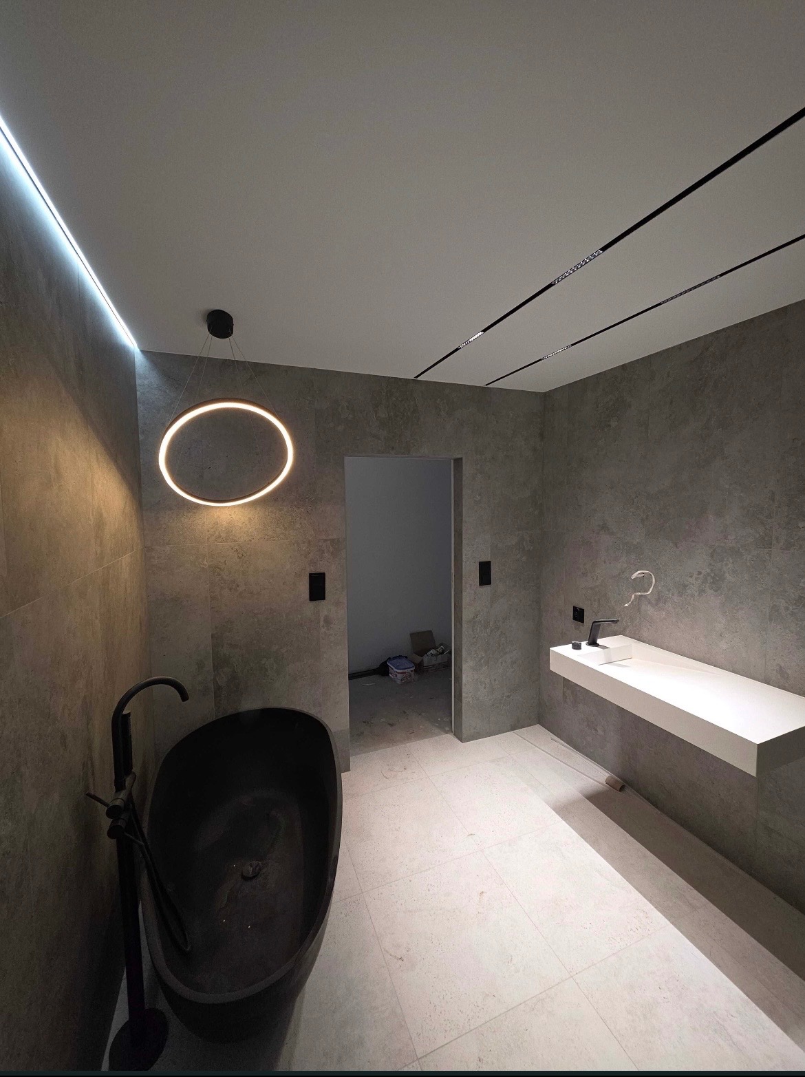 Nowoczesna łazienka z czarną wanną wolnostojącą i podświetlanym lustrem w kształcie okręgu. Szare płytki na ścianach i jasna podłoga. Minimalistyczny design.