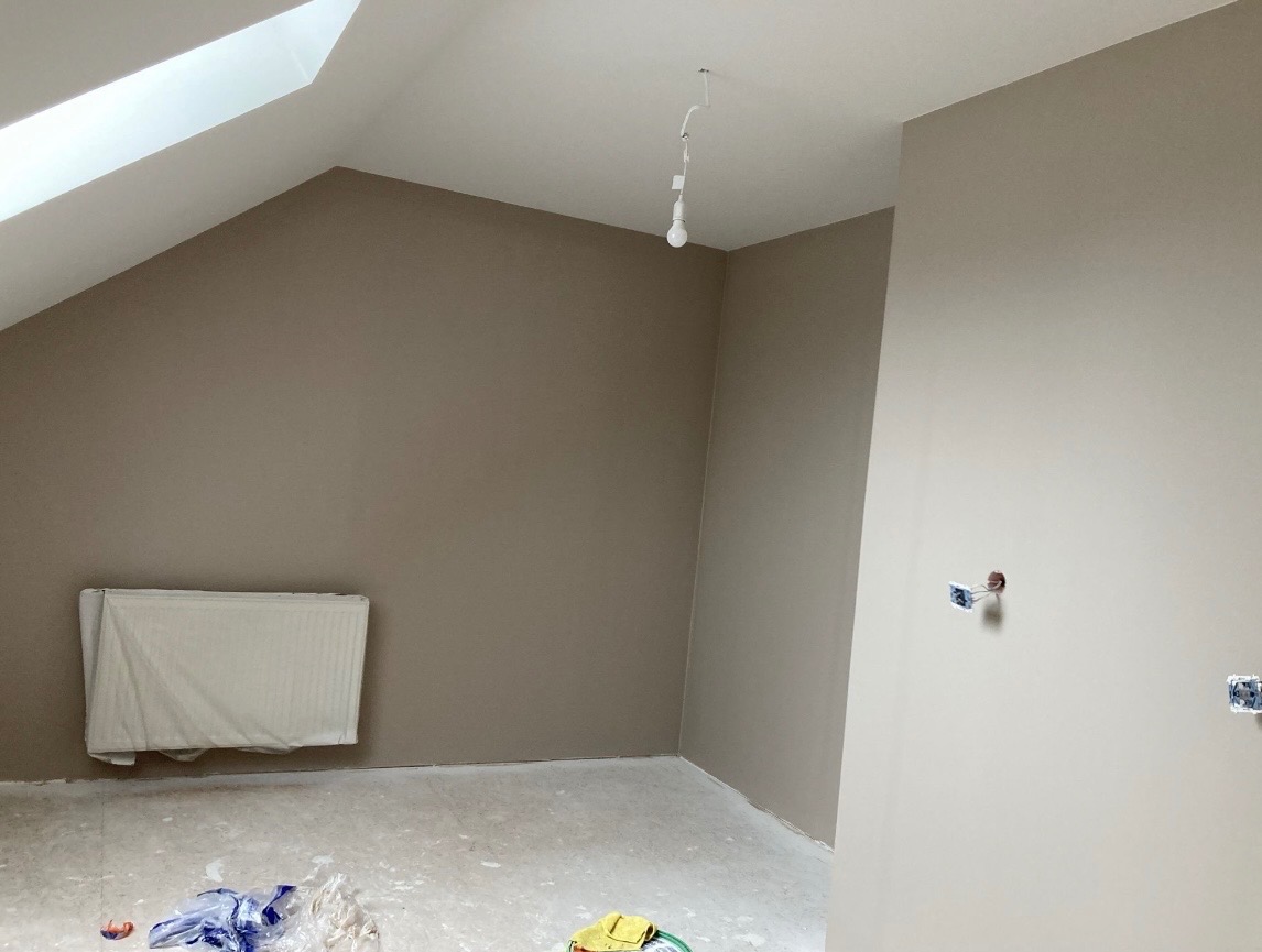 Pokój na poddaszu po malowaniu ścian na beżowo-szary kolor. Widoczne gniazdka elektryczne bez osprzętu, zakryty folią grzejnik i surowe podłoże.