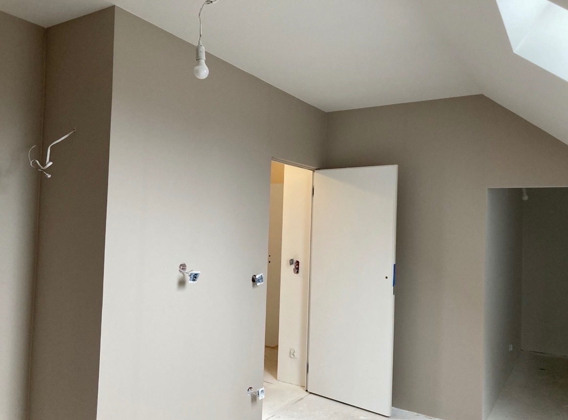 Wnętrze w trakcie malowania: ściany w odcieniach beżu, widoczne puszki elektryczne, surowa żarówka i białe drzwi. Skos dachu dodaje charakteru.