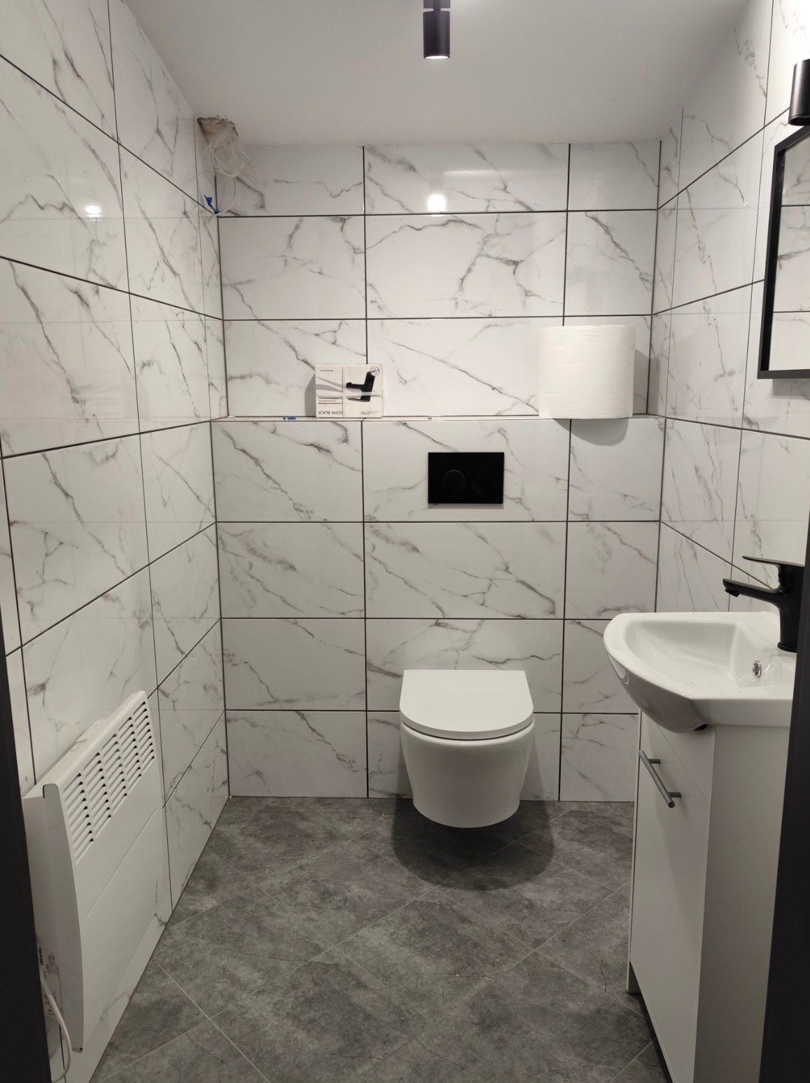 Nowoczesna łazienka po remoncie: białe płytki imitujące marmur, podwieszana toaleta, umywalka z szafką i grzejnik elektryczny. Minimalistyczny design, czarne akcenty.