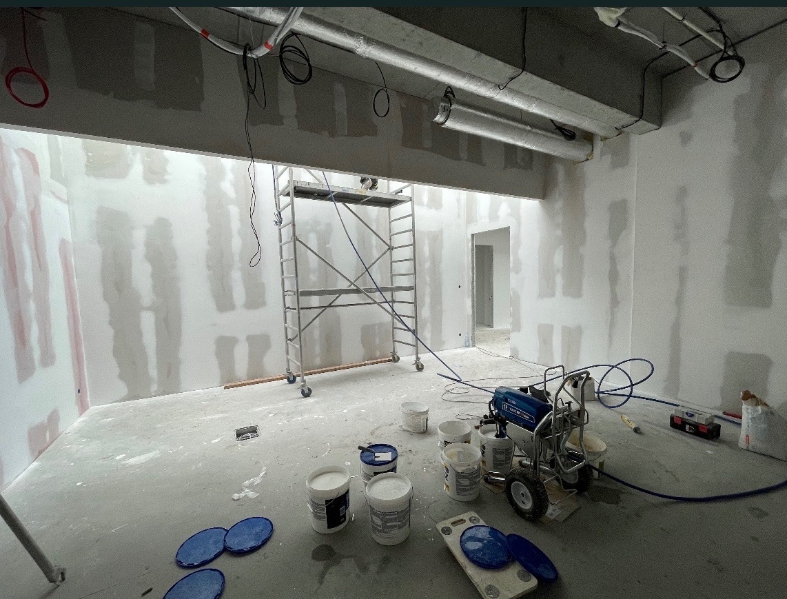 Wnętrze w trakcie szpachlowania maszynowego. Widoczne ściany z nałożoną szpachlą, sprzęt do natrysku, wiaderka z masą i rusztowanie. Przewody pod sufitem.