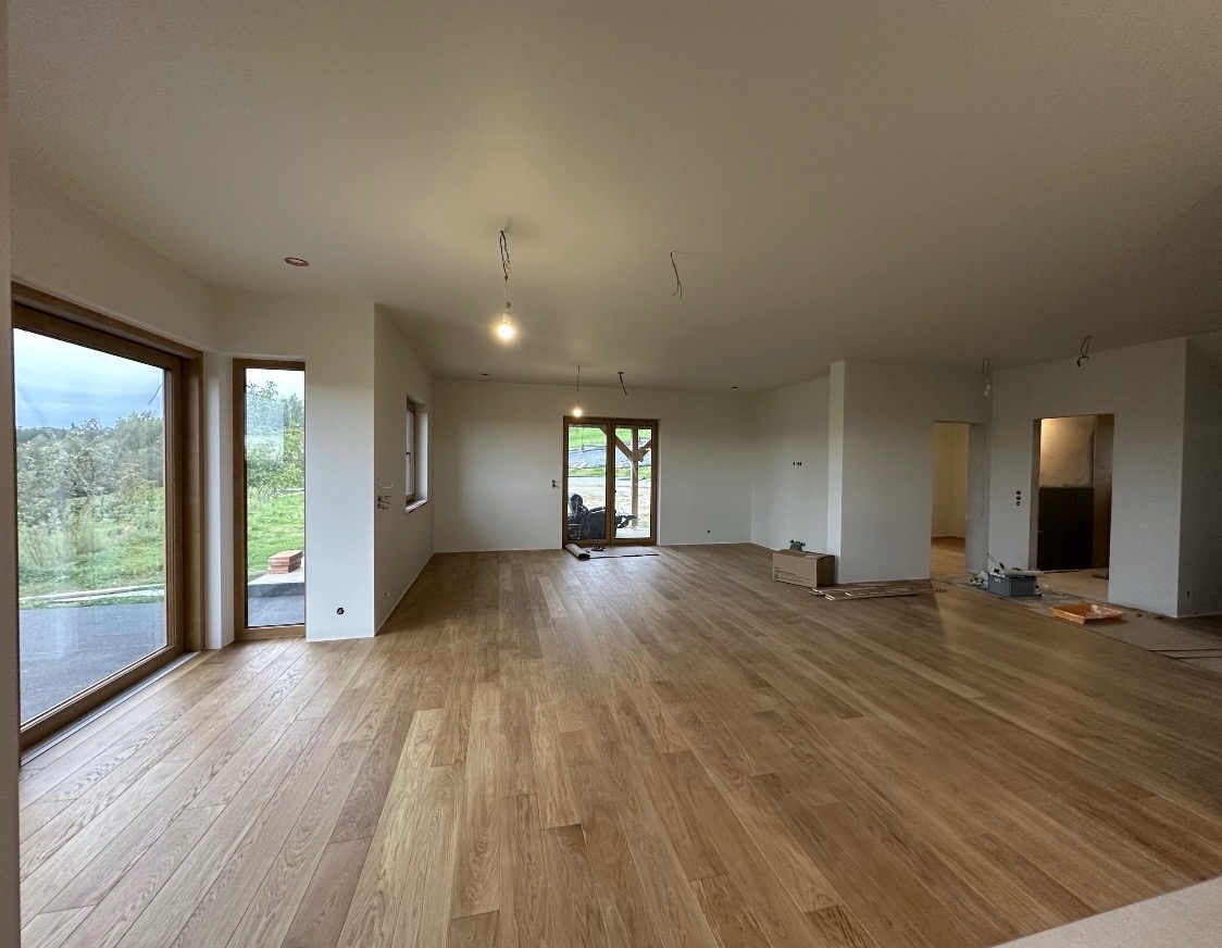 Jasne, przestronne wnętrze domu po remoncie: drewniana podłoga, białe ściany, duże okna z widokiem na zieleń. Widoczne przygotowania do montażu oświetlenia.