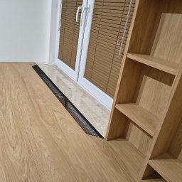 Franciszek Mętel - Biurko z drewnianym blatem i regałem, wkomponowane w przestrzeń okienną z żaluzjami. Nowoczesny design, jasne drewno, czarna siatka wentylacyjna.