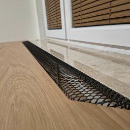 Franciszek Mętel - Szczegółowe ujęcie podłogi z jasnego drewna i czarnej, metalowej kratki wentylacyjnej przy oknie z drewnianymi żaluzjami. Minimalistyczny design.