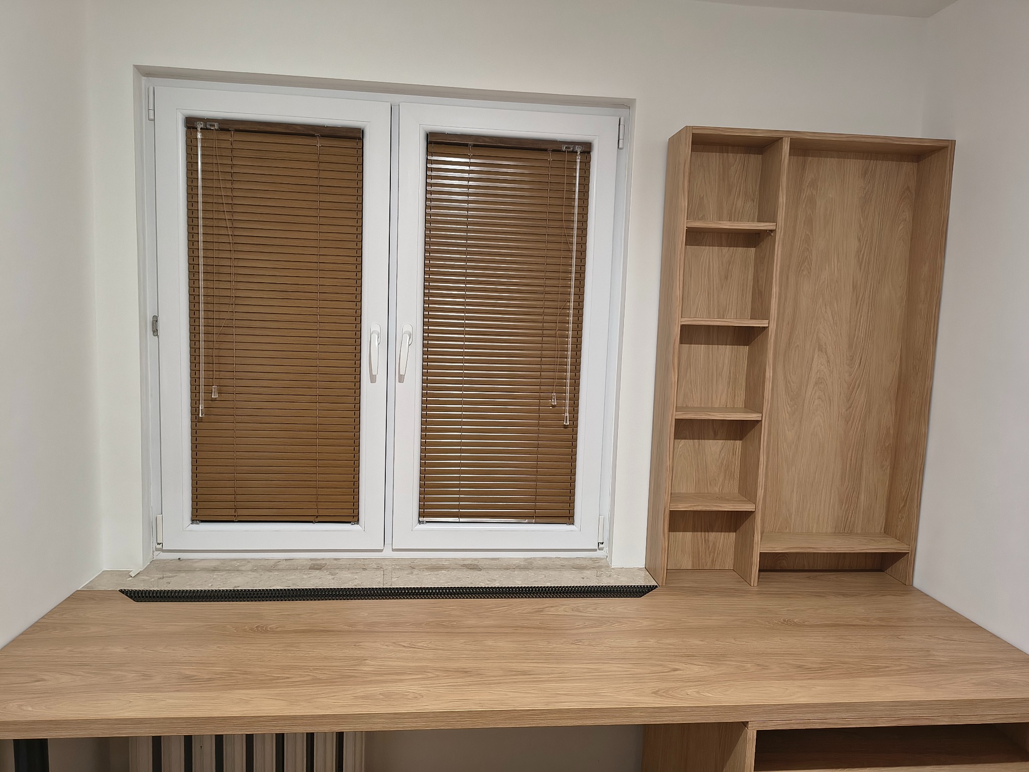 Drewniane biurko z regałem wnękowym przy oknie z żaluzjami. Jasne drewno, minimalistyczny design, zabudowa na wymiar, widok z przodu, oświetlenie naturalne.