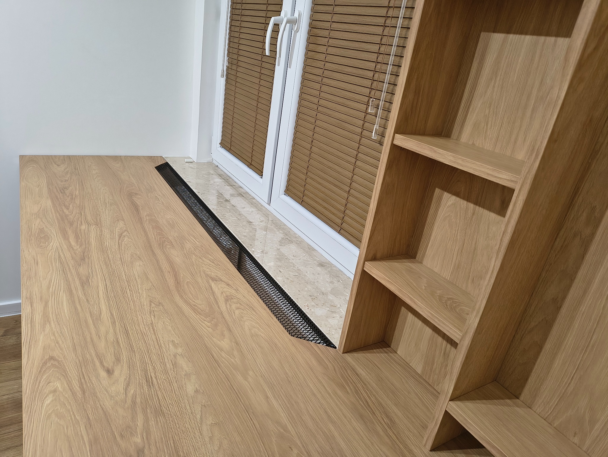 Biurko z drewnianym blatem i regałem, wkomponowane w przestrzeń okienną z żaluzjami. Nowoczesny design, jasne drewno, czarna siatka wentylacyjna.