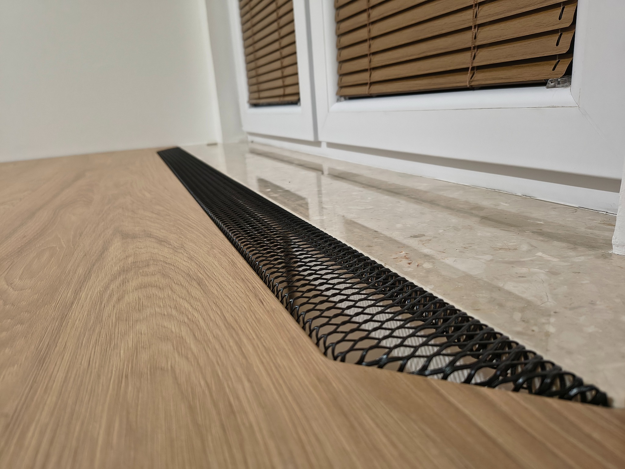 Szczegółowe ujęcie podłogi z jasnego drewna i czarnej, metalowej kratki wentylacyjnej przy oknie z drewnianymi żaluzjami. Minimalistyczny design.