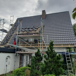 VIS - Dom w trakcie krycia dachu czarną dachówką. Widoczne rusztowania, częściowo ułożone dachówki i drewniana konstrukcja. Pochmurne niebo w tle.