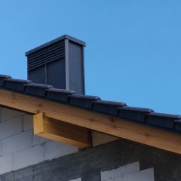 VIS - Częściowy widok dachu z czarną dachówką, drewnianą więźbą i kominem wentylacyjnym na tle błękitnego nieba. Widoczny fragment ściany z białych bloczków.