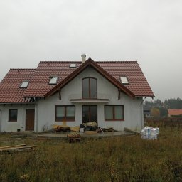 VIS - Dom jednorodzinny z czerwoną dachówką, widoczne okna dachowe i elementy wykończeniowe. Na pierwszym planie trawa i materiały budowlane. Pochmurne niebo w tle.