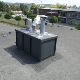 VIS - Szary, płaski dach budynku z zainstalowanymi elementami wentylacyjnymi i kominami. W tle budynek z napisem 'IO ARBOVEN'.