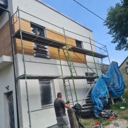 Ecoenergy 4 You - Dwukondygnacyjny dom z rusztowaniem podczas prac elewacyjnych. Część ściany pomalowana na biało, fragment wykończony drewnopodobną okładziną. Okna zabezpieczone folią.