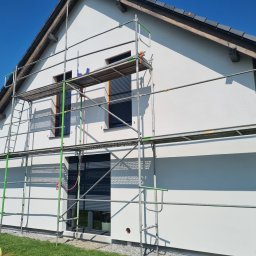 Ecoenergy 4 You - Świeżo otynkowana, biała elewacja domu jednorodzinnego w słoneczny dzień, z widocznym rusztowaniem i fragmentem zielonego trawnika. Okna z szarymi roletami.