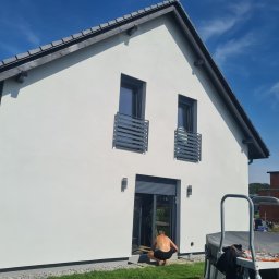 Ecoenergy 4 You - Biała elewacja domu jednorodzinnego z szarymi oknami i dachem. Osoba w kucki przy drzwiach tarasowych. Widoczne rusztowanie i fragment ogrodu. Słoneczny dzień.