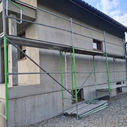 Ecoenergy 4 You - Dom jednorodzinny w trakcie tynkowania elewacji, widoczne rusztowania z elementami w kolorze zielonym. Świeży tynk o szarej barwie. Dom otoczony kamieniami.
