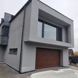 Ecoenergy 4 You - Nowoczesny dom z szarą elewacją i drewnianą bramą garażową. Część budynku wykończona kamieniem. Widoczne drzewa i fragment otoczenia.