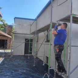 Ecoenergy 4 You - Pracownik na rusztowaniu, w trakcie prac elewacyjnych na tle budynku z szarą elewacją. Widoczne zabezpieczenia na ziemi i fragmenty sąsiednich budynków.