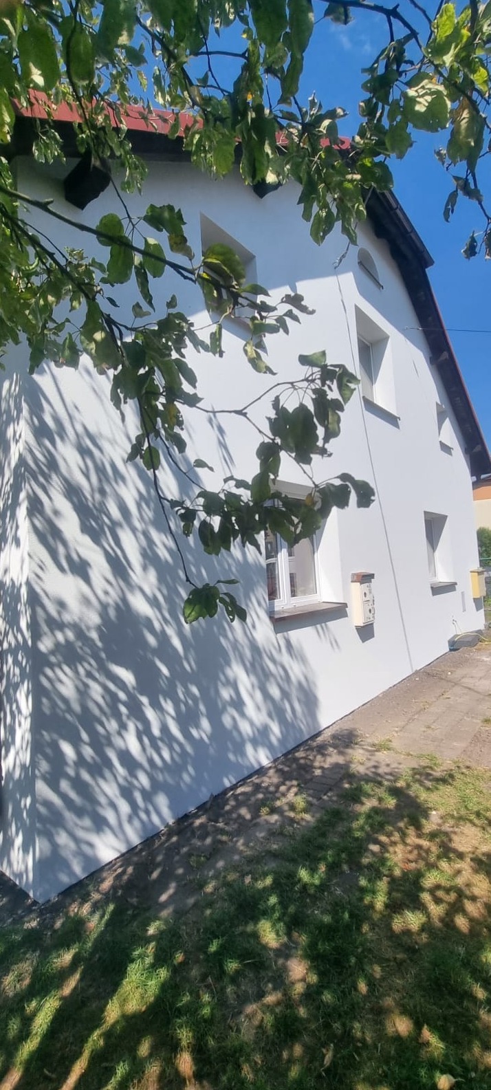 Boczny widok domu z białą elewacją, częściowo zacienioną przez liście drzew. Widoczne okna, skrzynka pocztowa i fragment dachu. Słoneczny dzień.