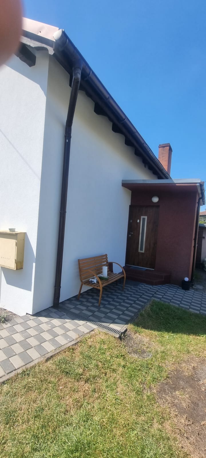 Biała elewacja domu jednorodzinnego z brązowymi rynnami i wejściem. Na chodniku stoi drewniana ławka z akcesoriami malarskimi. Błękitne niebo w tle.