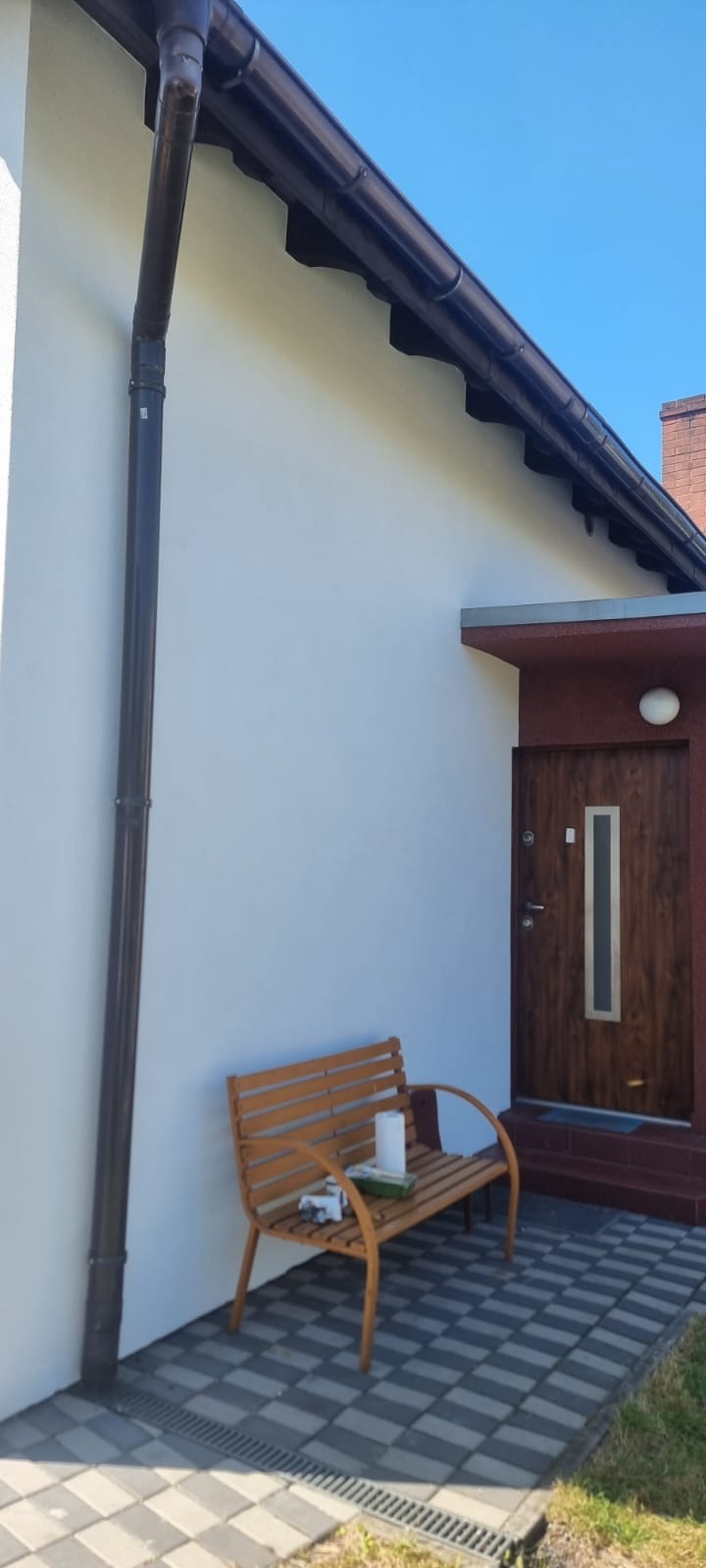 Biała elewacja domu jednorodzinnego z brązowymi rynnami i wejściem. Na pierwszym planie drewniana ławka z akcesoriami malarskimi. Słoneczny dzień.