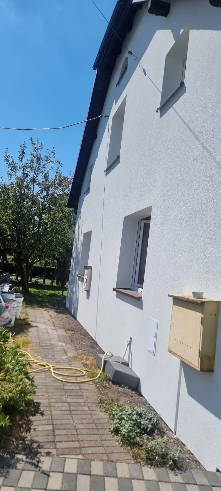 Biała elewacja domu jednorodzinnego z widocznymi oknami i skrzynką pocztową, sfotografowana w słoneczny dzień z perspektywy ścieżki.