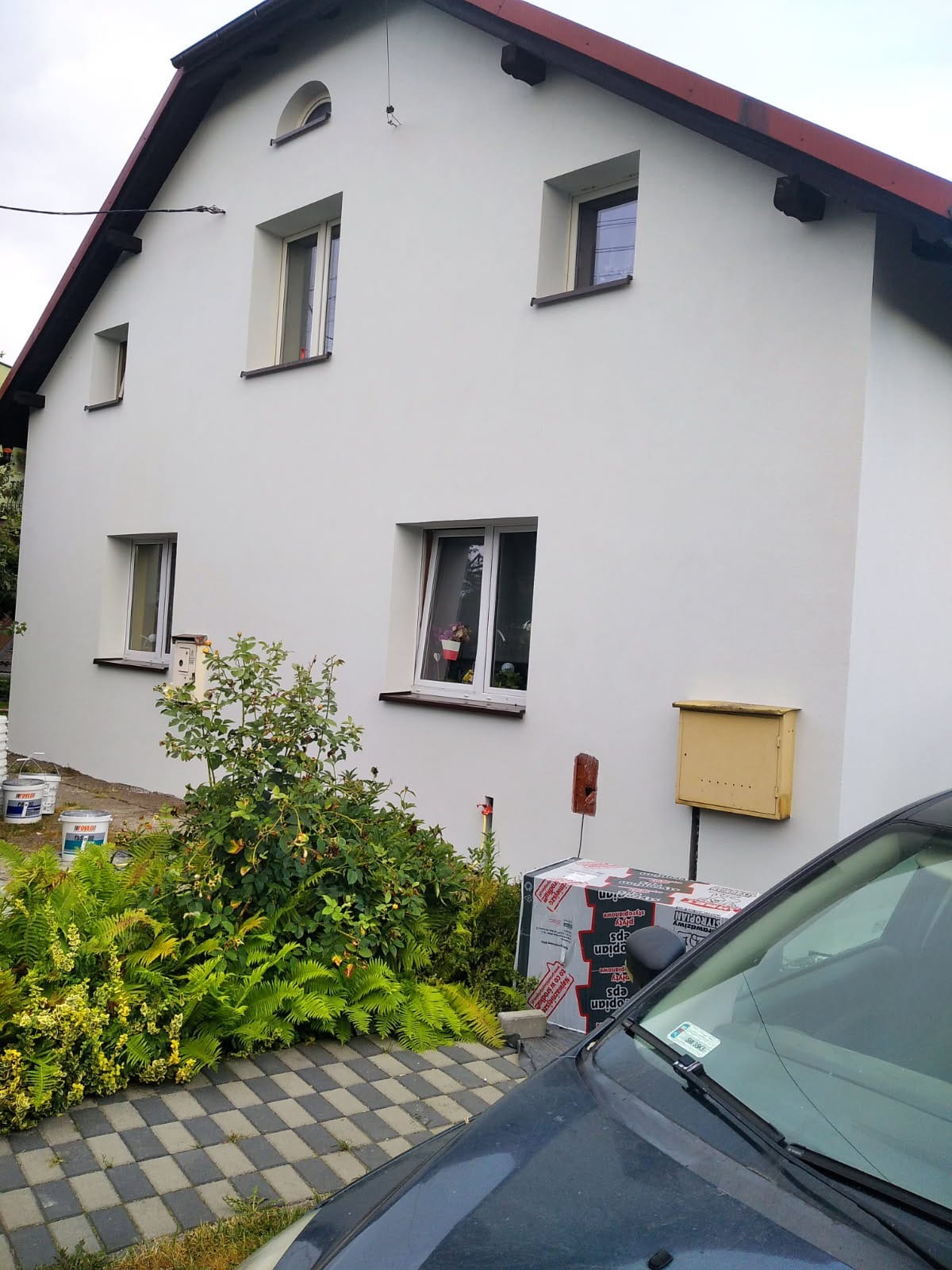 Biała elewacja domu jednorodzinnego z czerwonym dachem, widok z poziomu gruntu, z widocznymi puszkami farb i paczką styropianu w ogrodzie.