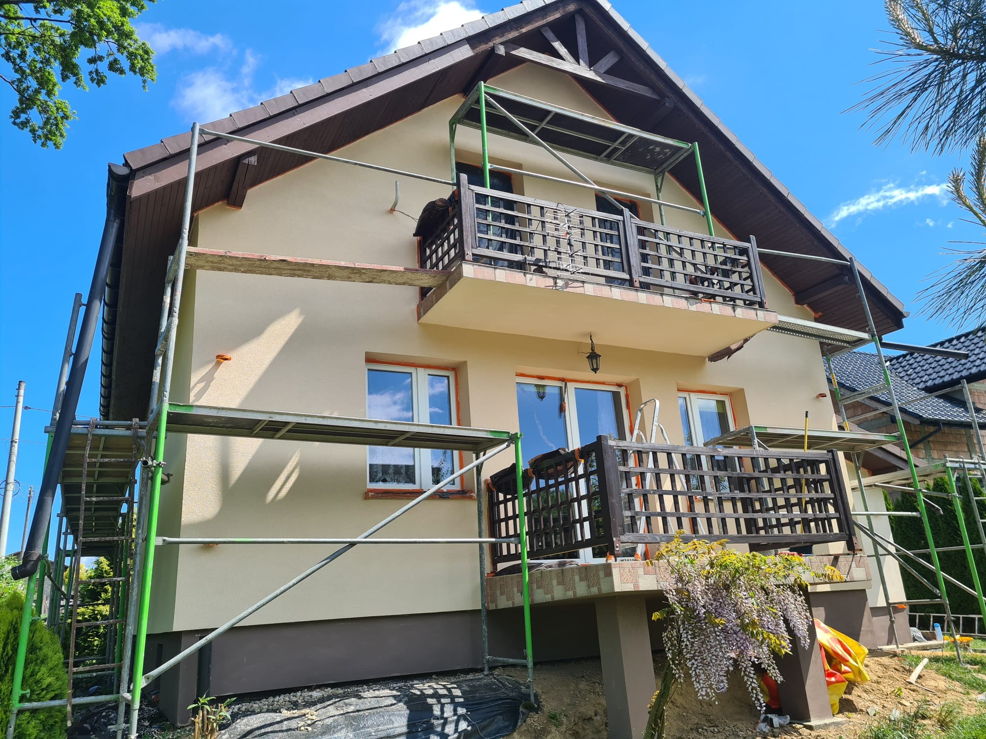 Dwupiętrowy dom w trakcie prac elewacyjnych, z rusztowaniem i widocznym balkonem z drewnianą balustradą. Świeżo otynkowana fasada w odcieniach beżu i brązu.