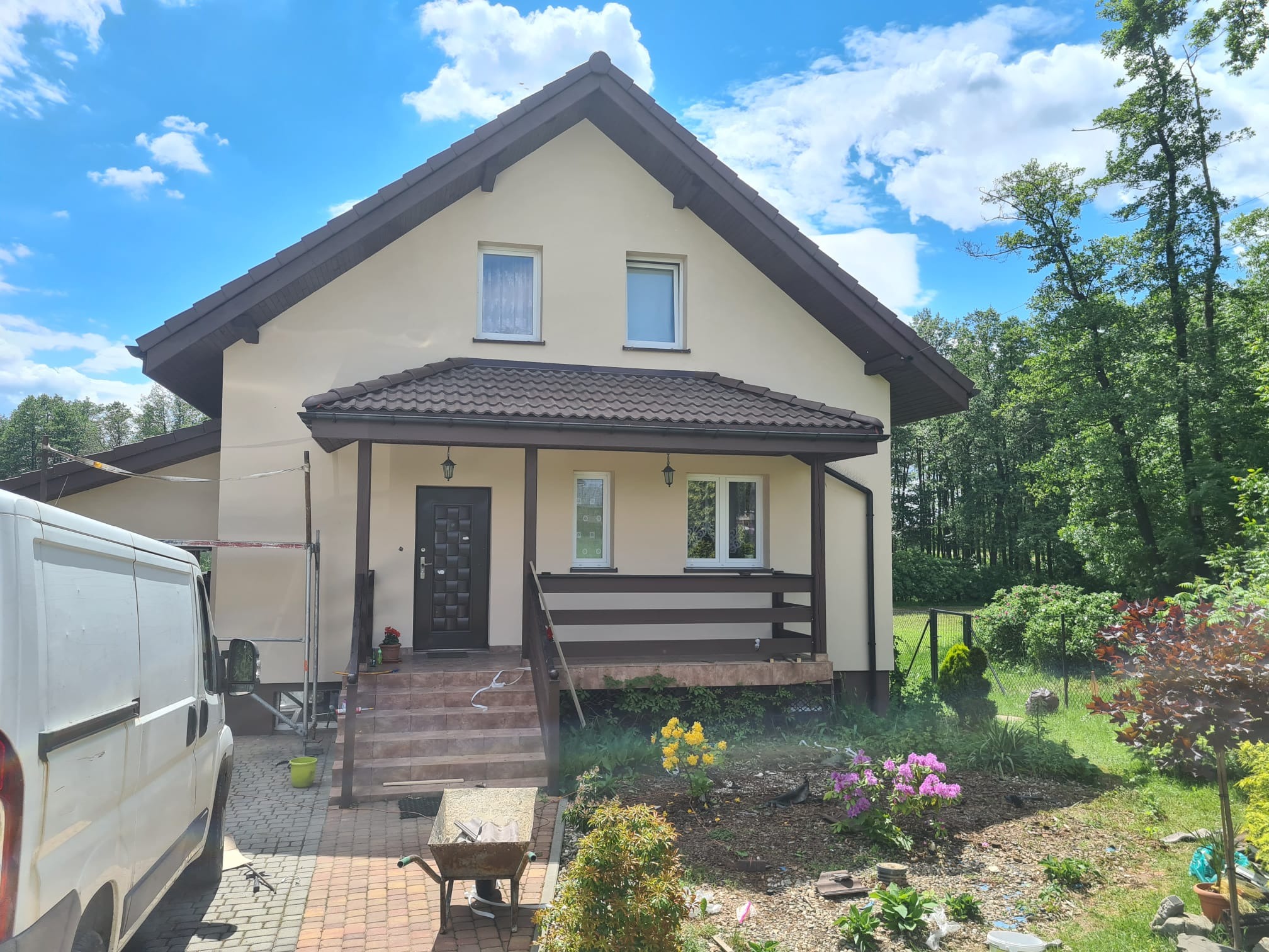 Jasna elewacja domu jednorodzinnego z brązowym dachem i werandą. Na pierwszym planie taczka i częściowo widoczny samochód dostawczy. Błękitne niebo z chmurami w tle.