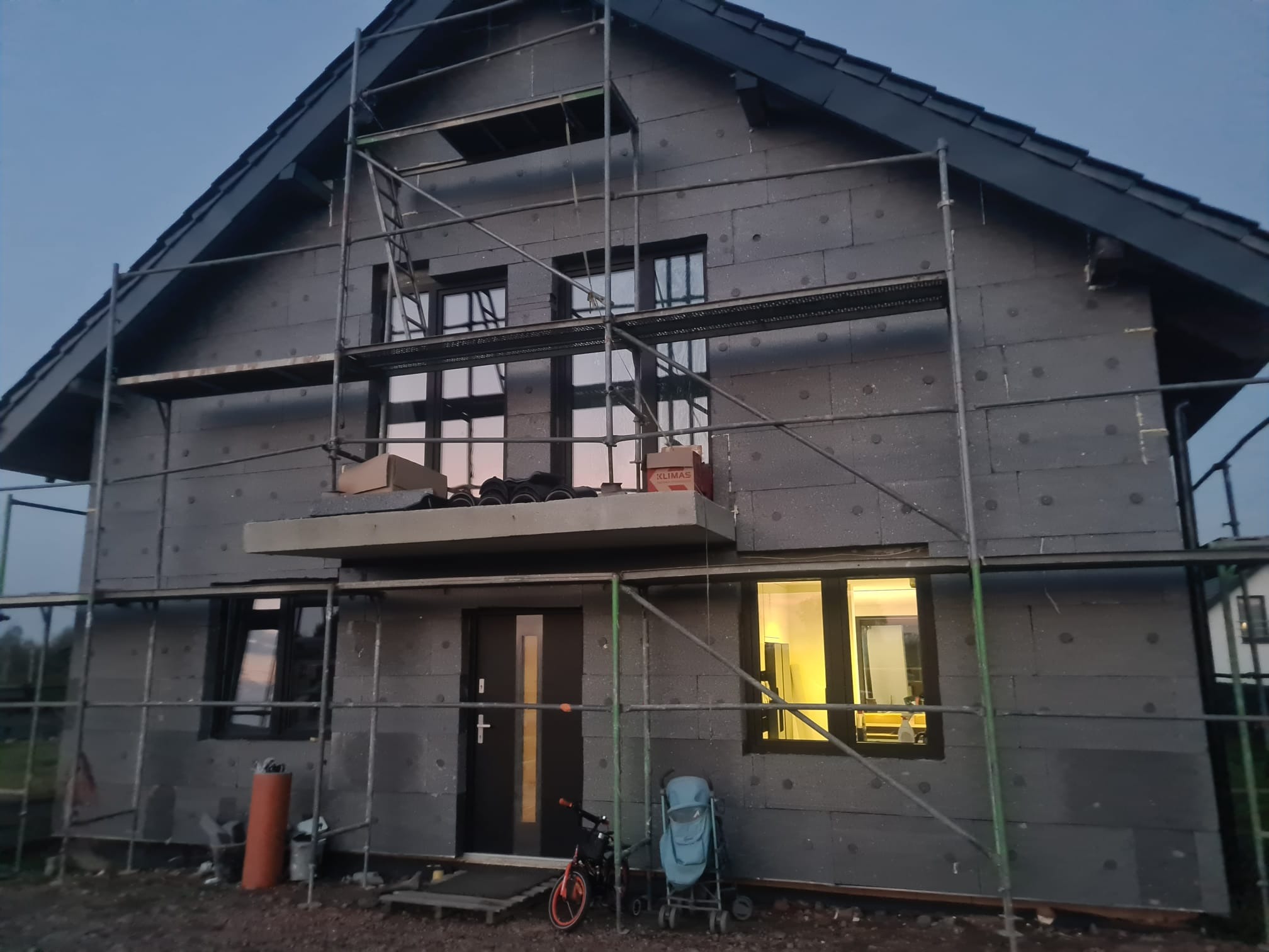 Dom jednorodzinny w trakcie ocieplania, widoczne rusztowania, szare płyty izolacyjne, okna z ciemnymi ramami. Na pierwszym planie dziecięcy wózek i rowerek.