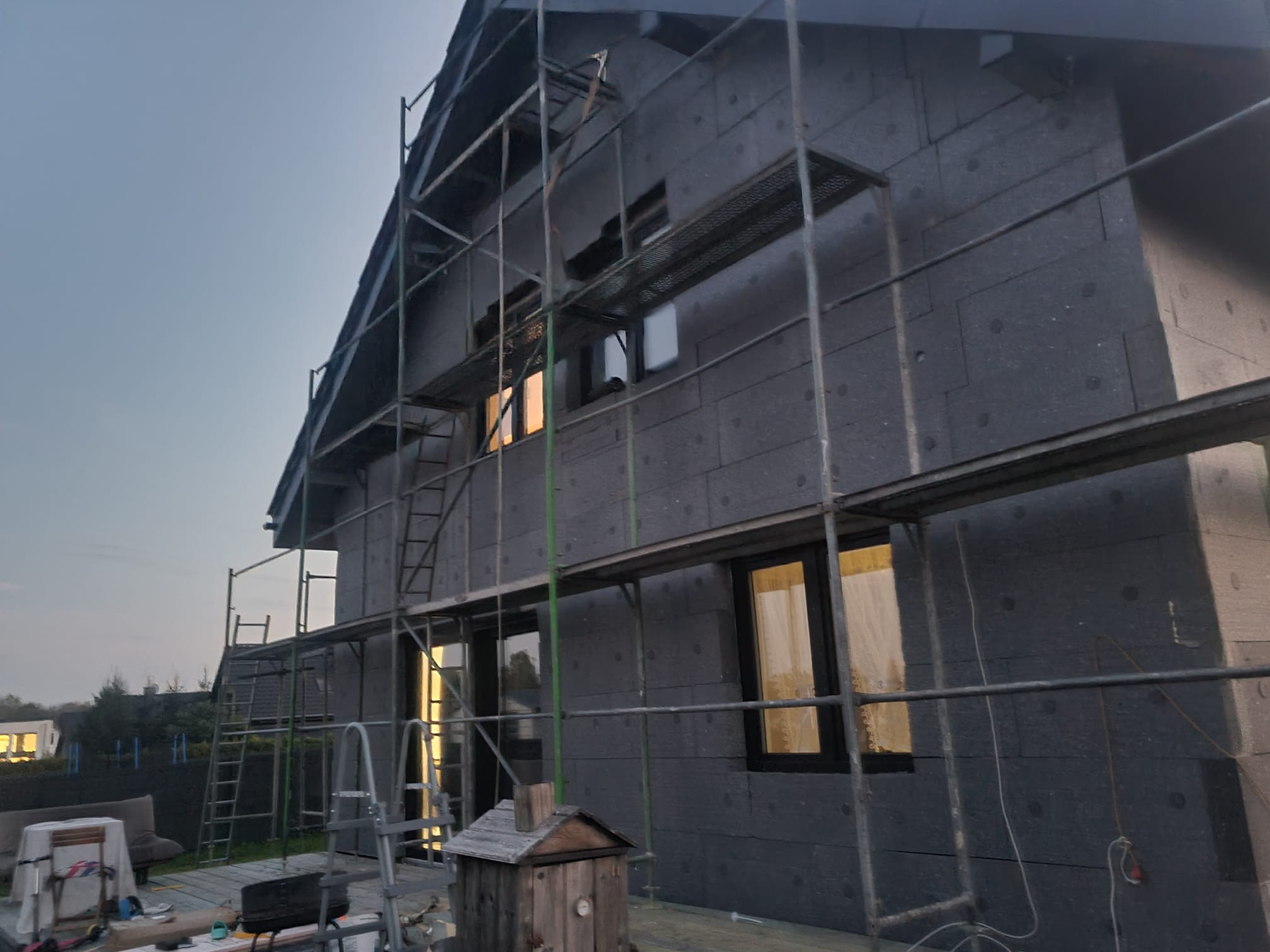 Dom w trakcie ocieplania, widoczne rusztowania i ciemna warstwa izolacyjna na elewacji. Okna zabezpieczone folią. Prace w toku, widok z zewnątrz.