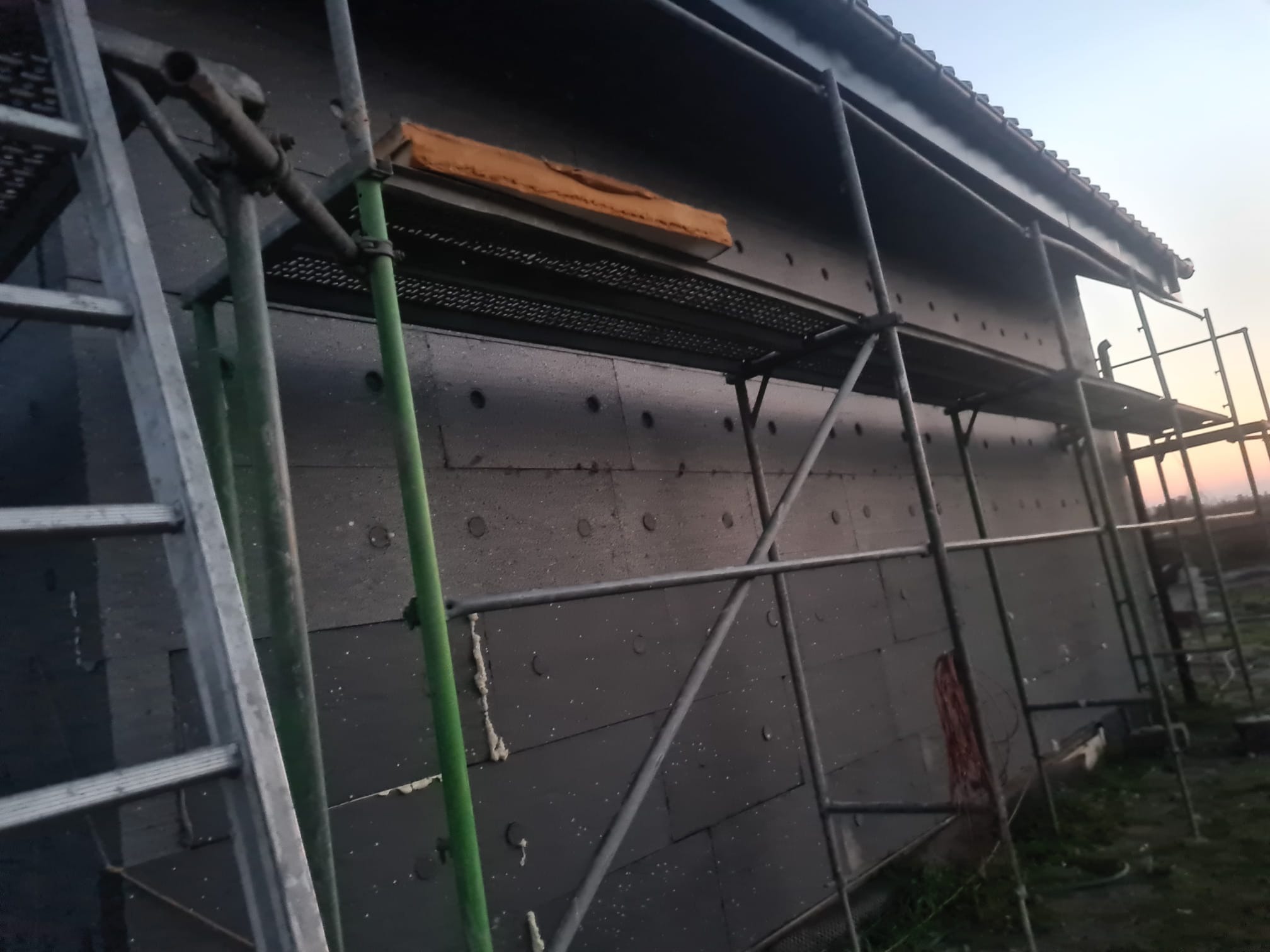 Dom w trakcie ocieplania, widoczne rusztowania i płyty izolacyjne na ścianie. Fragment dachu i nieba w tle. Prace w toku, widoczne detale montażu.