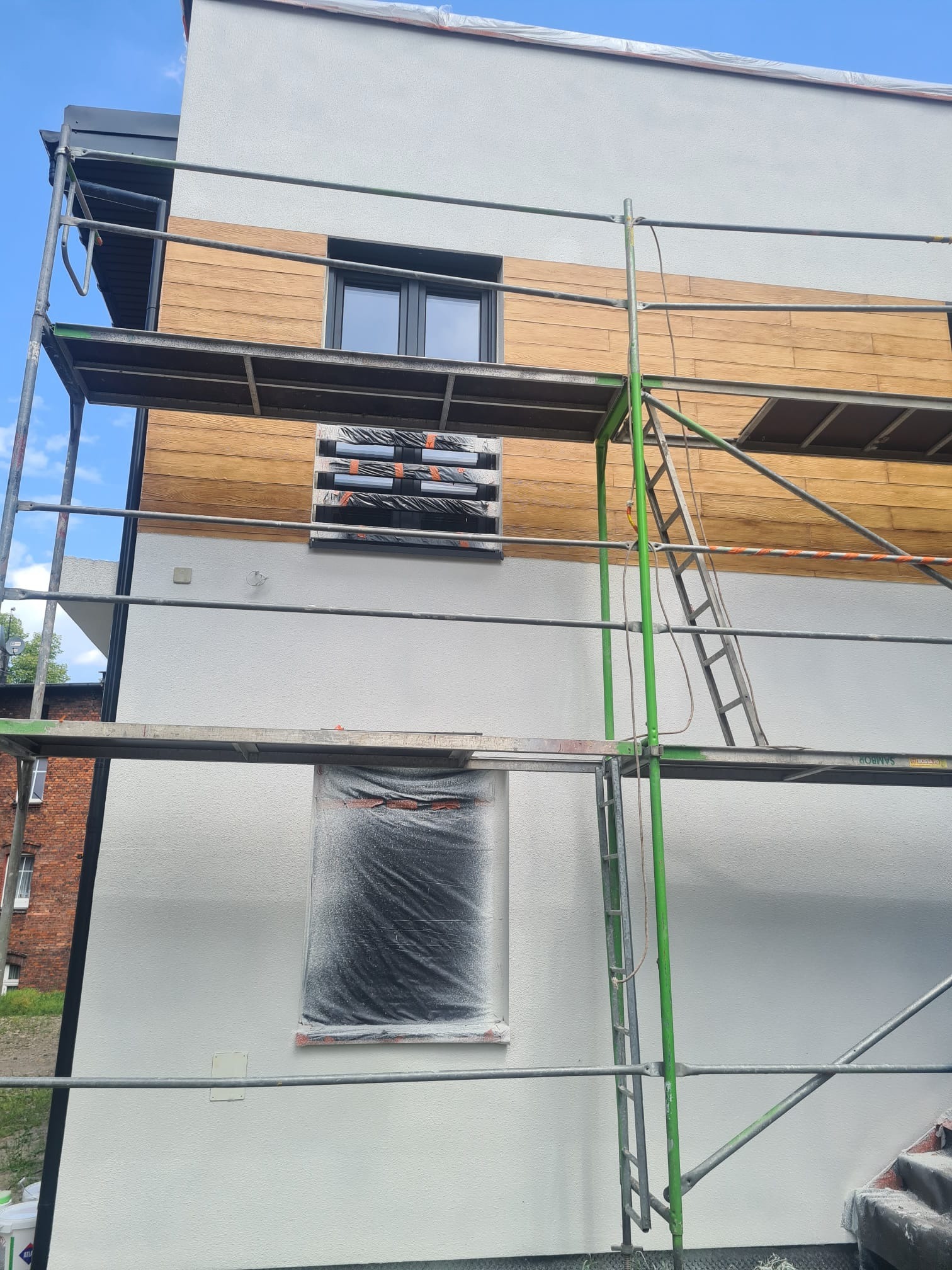 Dom w trakcie prac elewacyjnych: biały tynk, drewnopodobna okładzina, rusztowania. Okna zabezpieczone folią. Częściowo ukończona fasada.