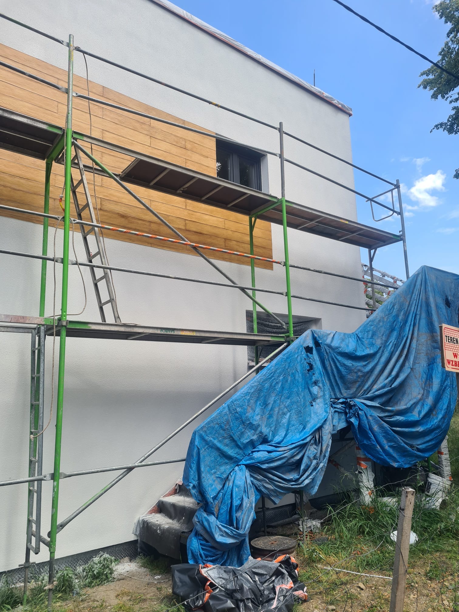 Elewacja domu w trakcie wykańczania z drewnianą okładziną, widoczne rusztowania i plandeka zabezpieczająca. Częściowo otynkowana ściana.