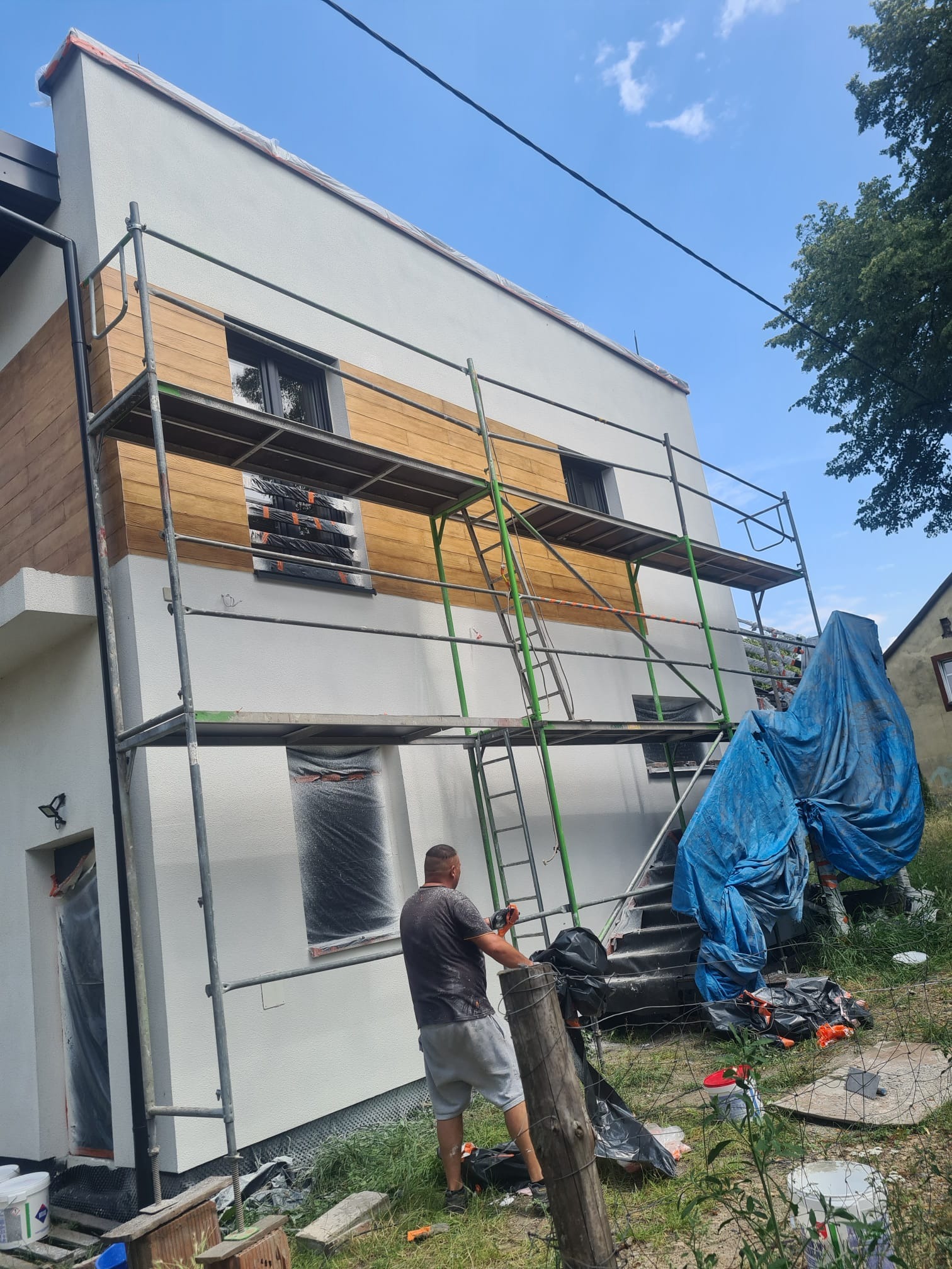 Dwukondygnacyjny dom z rusztowaniem podczas prac elewacyjnych. Część ściany pomalowana na biało, fragment wykończony drewnopodobną okładziną. Okna zabezpieczone folią.