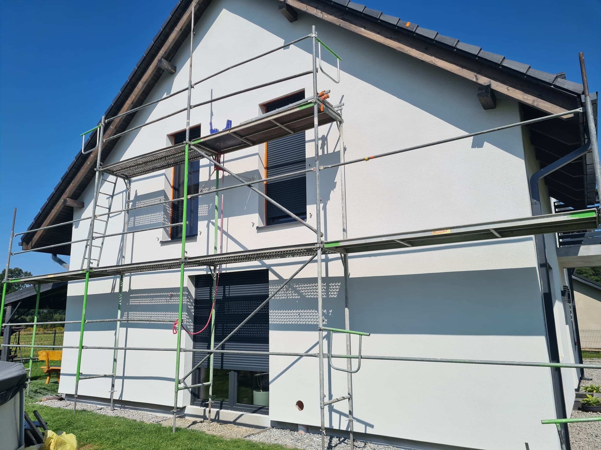 Świeżo otynkowana, biała elewacja domu jednorodzinnego w słoneczny dzień, z widocznym rusztowaniem i fragmentem zielonego trawnika. Okna z szarymi roletami.