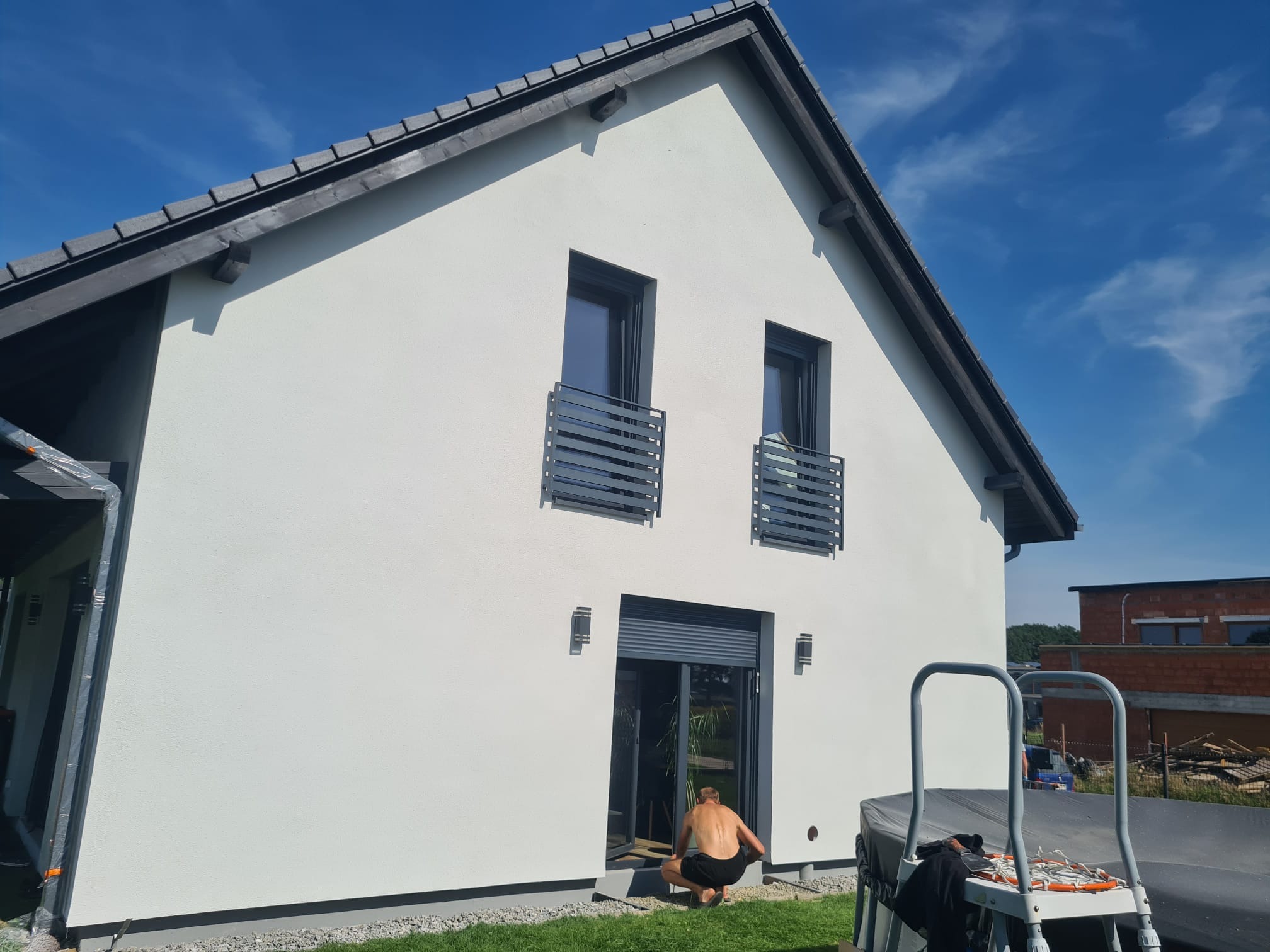 Biała elewacja domu jednorodzinnego z szarymi oknami i dachem. Osoba w kucki przy drzwiach tarasowych. Widoczne rusztowanie i fragment ogrodu. Słoneczny dzień.