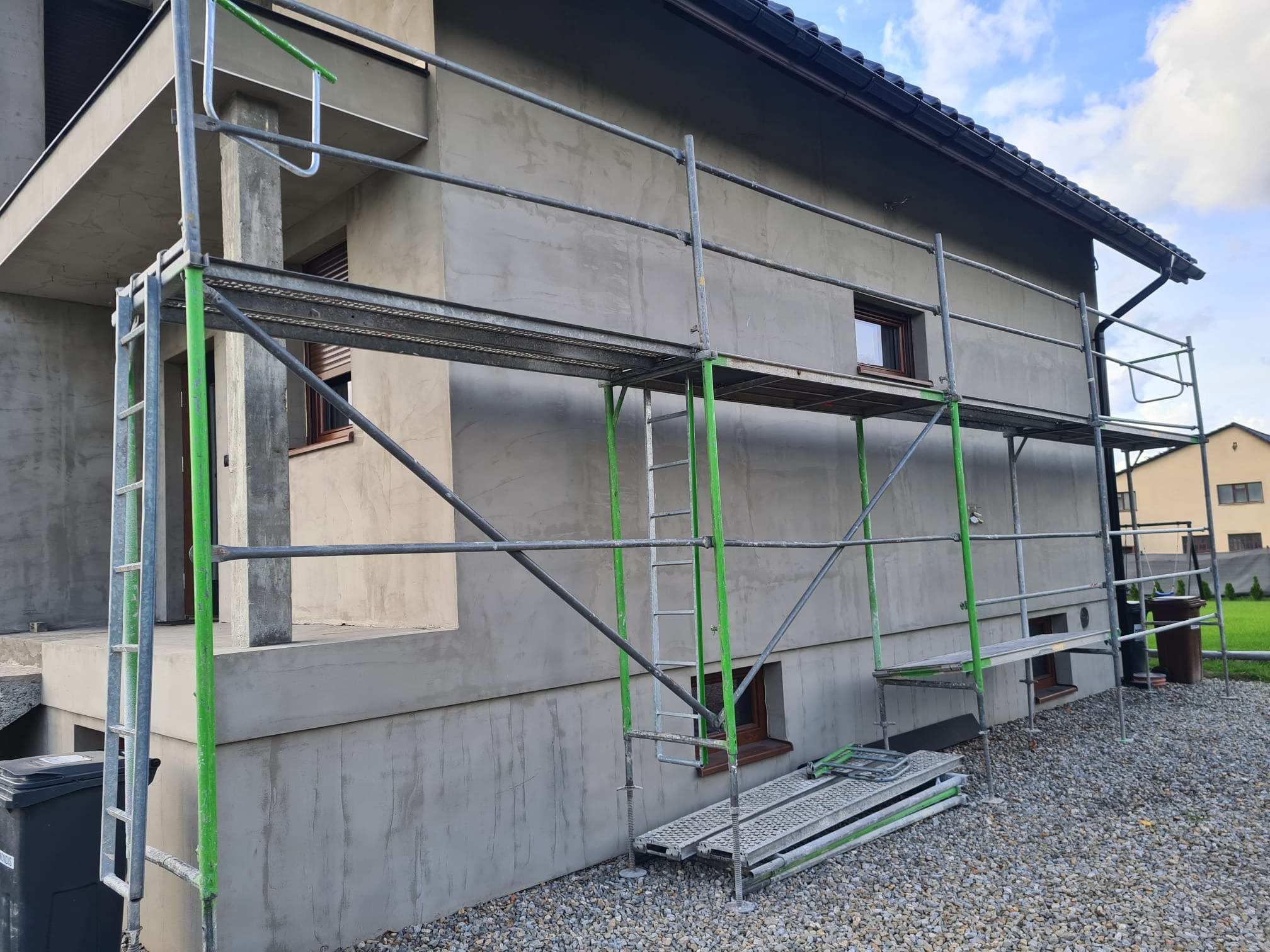 Dom jednorodzinny w trakcie tynkowania elewacji, widoczne rusztowania z elementami w kolorze zielonym. Świeży tynk o szarej barwie. Dom otoczony kamieniami.