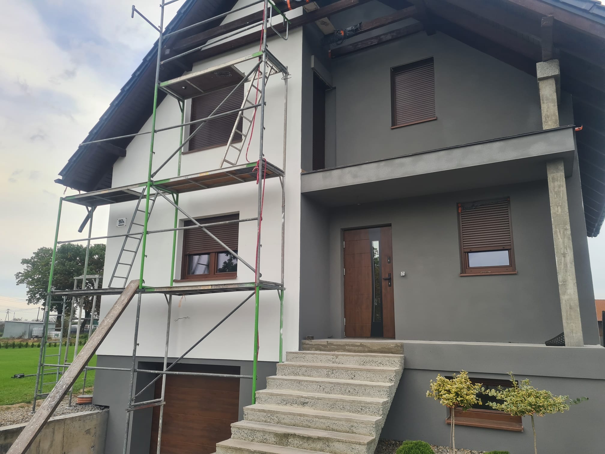 Dwukolorowy dom z rusztowaniem przy białej ścianie. Szara elewacja z brązowymi roletami i drzwiami. Schody prowadzą do wejścia. Widok na trawnik i drzewa.