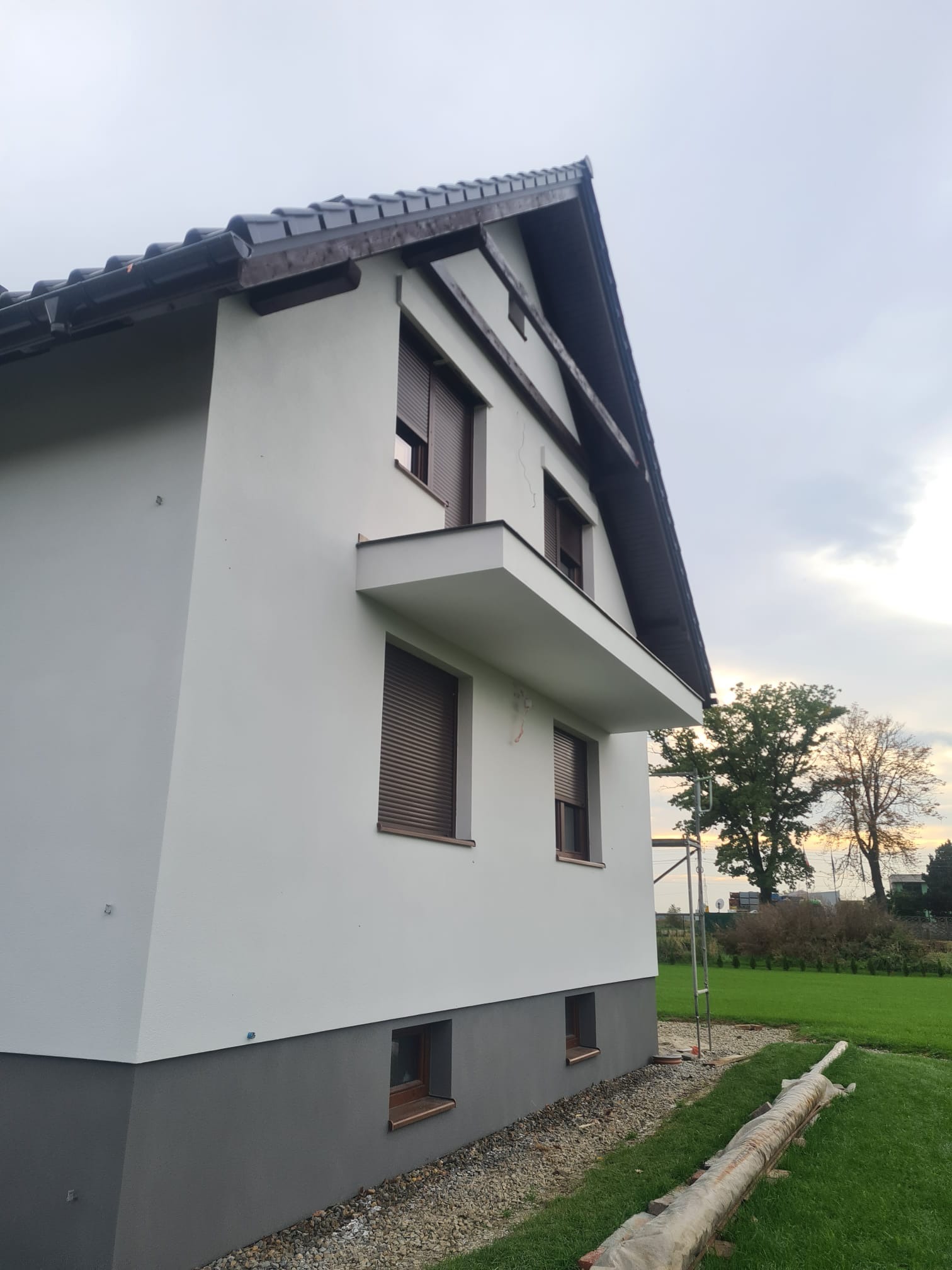 Boczny widok domu po malowaniu elewacji na biało z szarym cokołem. Widoczne okna z brązowymi roletami, balkon i fragment rusztowania.