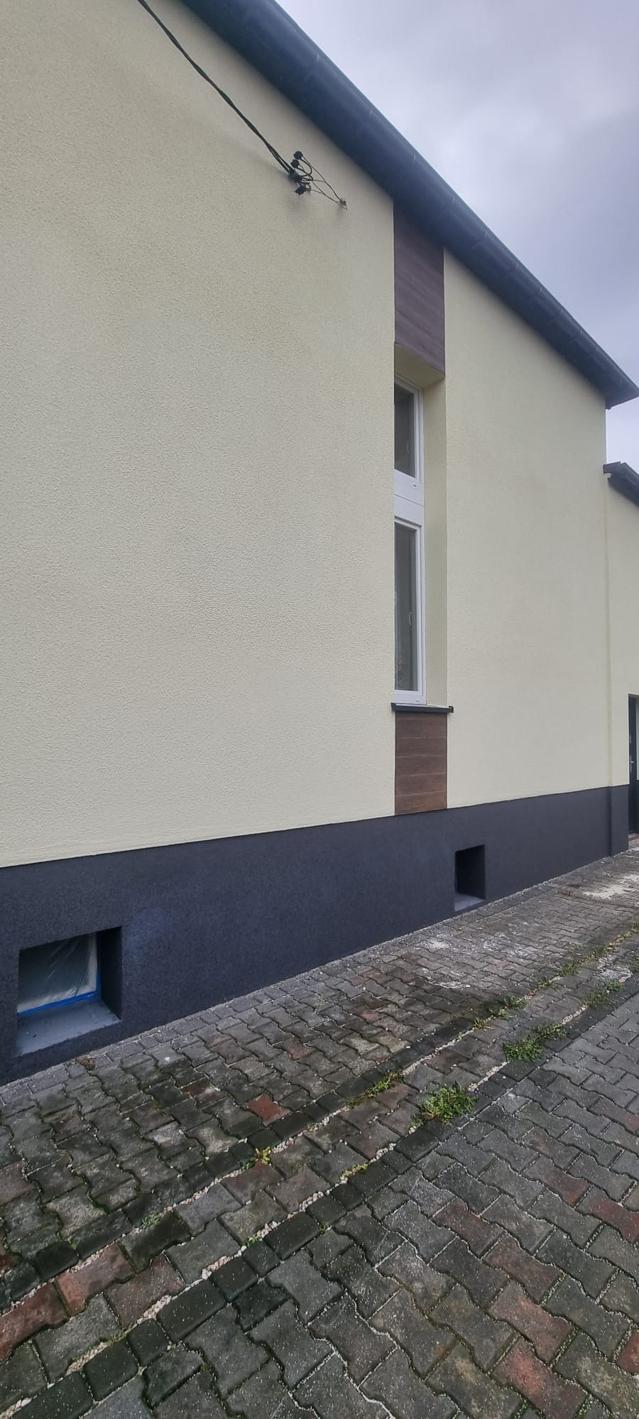 Jasna elewacja budynku z ciemną podmurówką i drewnopodobnymi elementami dekoracyjnymi. Widoczne okno i kratka wentylacyjna. Kostka brukowa na pierwszym planie.