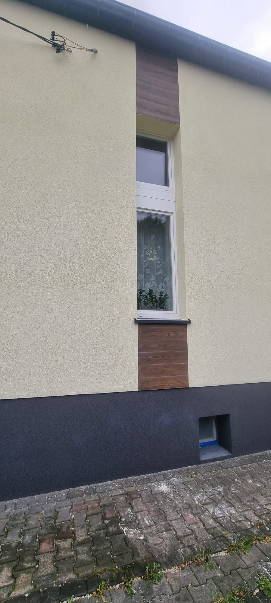 Elewacja budynku z oknem, wykończona tynkiem w odcieniach beżu i szarości, z dekoracyjnymi panelami imitującymi drewno wokół okna. Widoczny fragment chodnika.