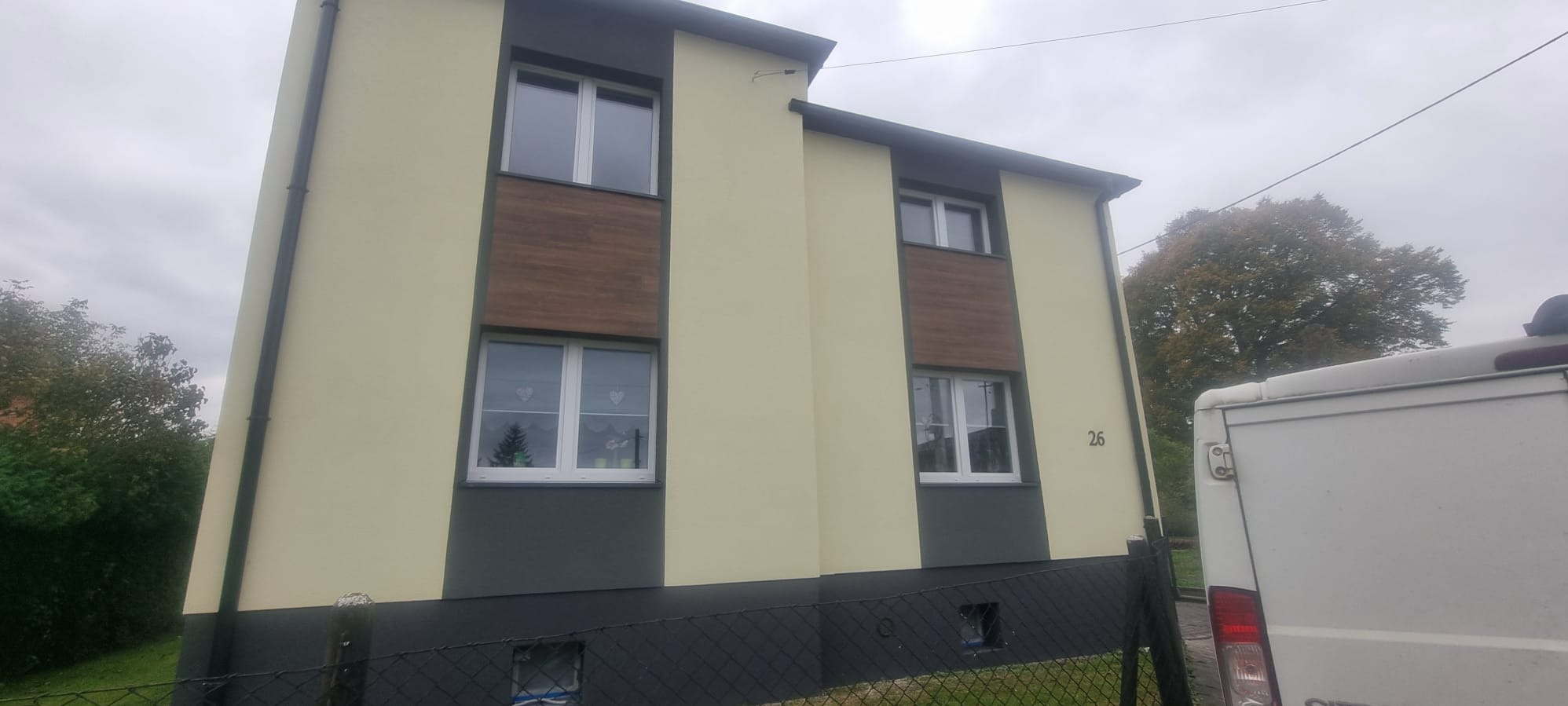 Dwukondygnacyjny dom z elewacją w odcieniach beżu i szarości, z drewnopodobnymi panelami między oknami. Widoczny fragment ogrodzenia i samochód dostawczy.