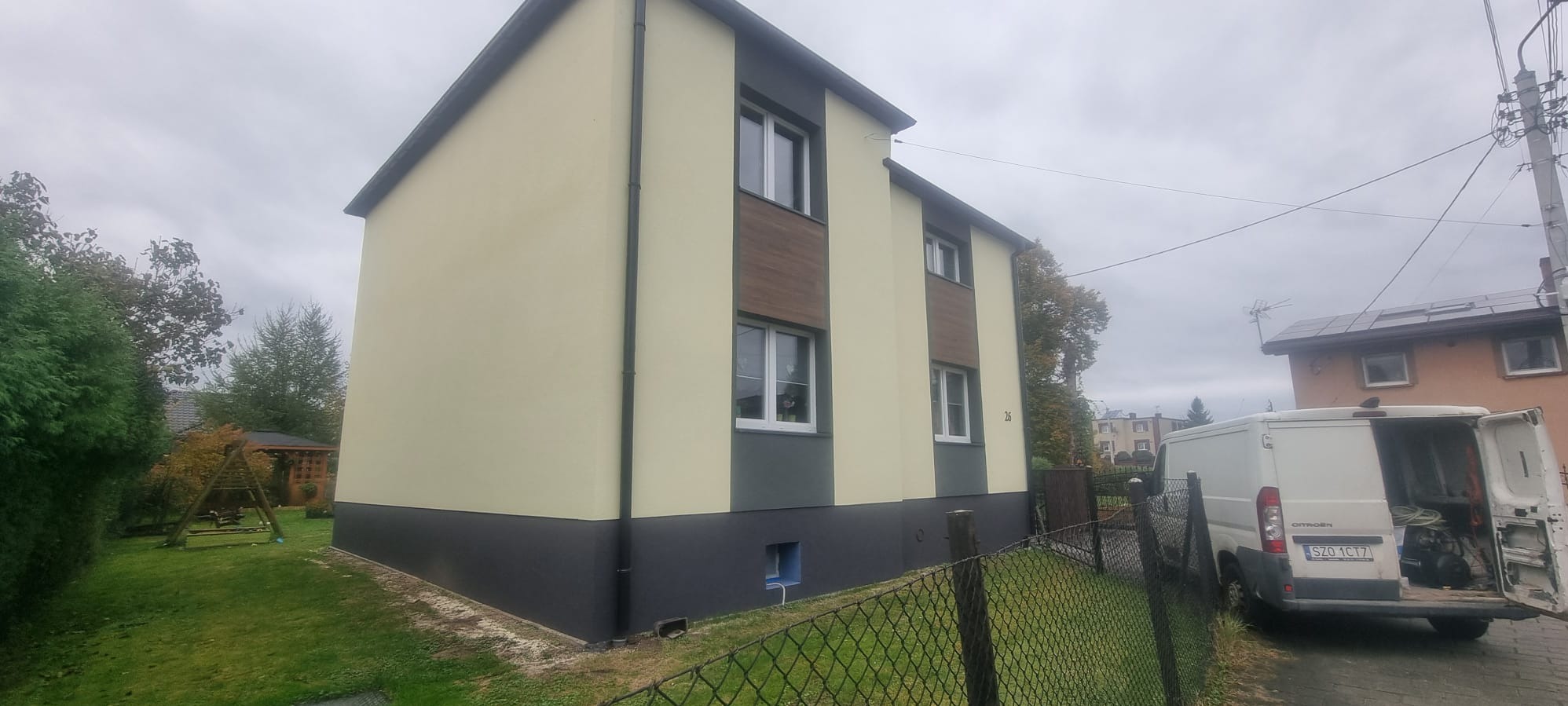 Dwukondygnacyjny dom po renowacji elewacji w odcieniach beżu i szarości, z elementami dekoracyjnymi imitującymi drewno. Na posesji zaparkowany bus.