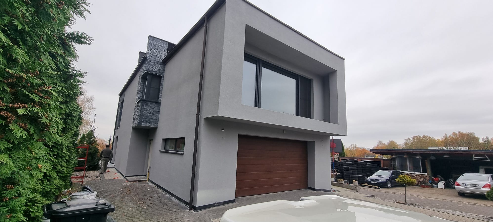 Nowoczesny dom z szarą elewacją i drewnianą bramą garażową. Część budynku wykończona kamieniem. Widoczne drzewa i fragment otoczenia.