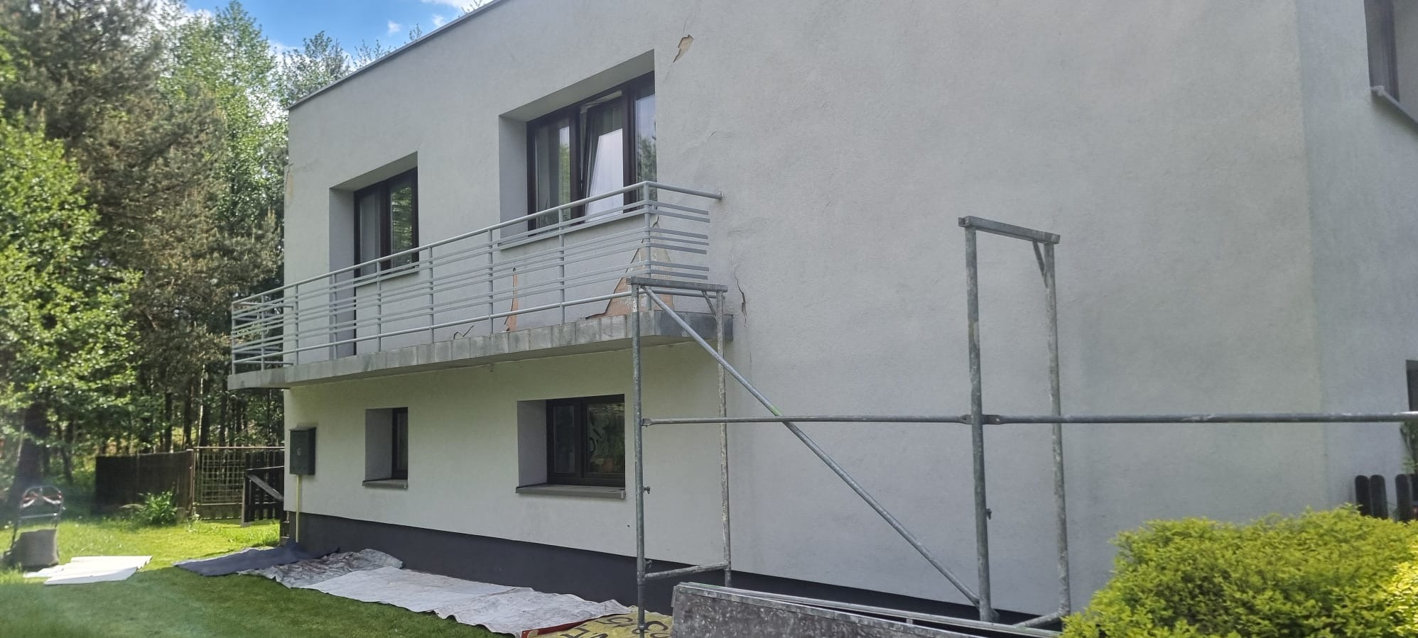 Dwupiętrowy dom z balkonem w trakcie prac elewacyjnych. Widoczne rusztowania i uszkodzenia tynku. Tło: zieleń drzew i trawnik.