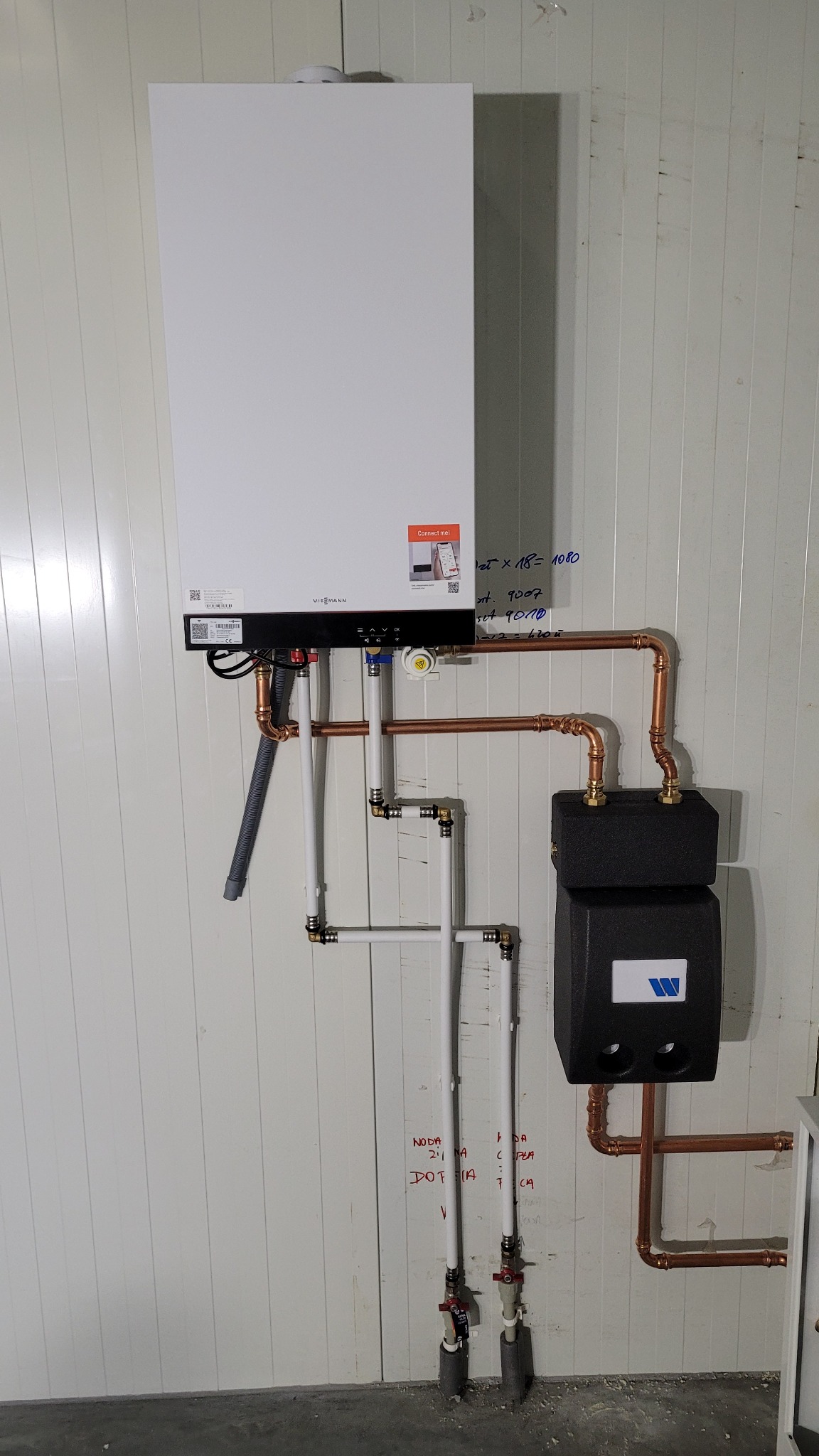 Biały piec gazowy Viessmann zamontowany na ścianie z panelami, z widocznymi miedzianymi i białymi rurami instalacji. Na ścianie notatki wykonane markerem.