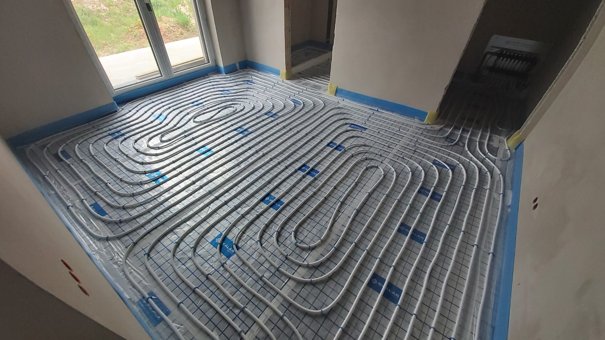 Białe rury ogrzewania podłogowego ułożone spiralnie na folii izolacyjnej z niebieskimi brzegami w surowym wnętrzu. Widoczny rozdzielacz.