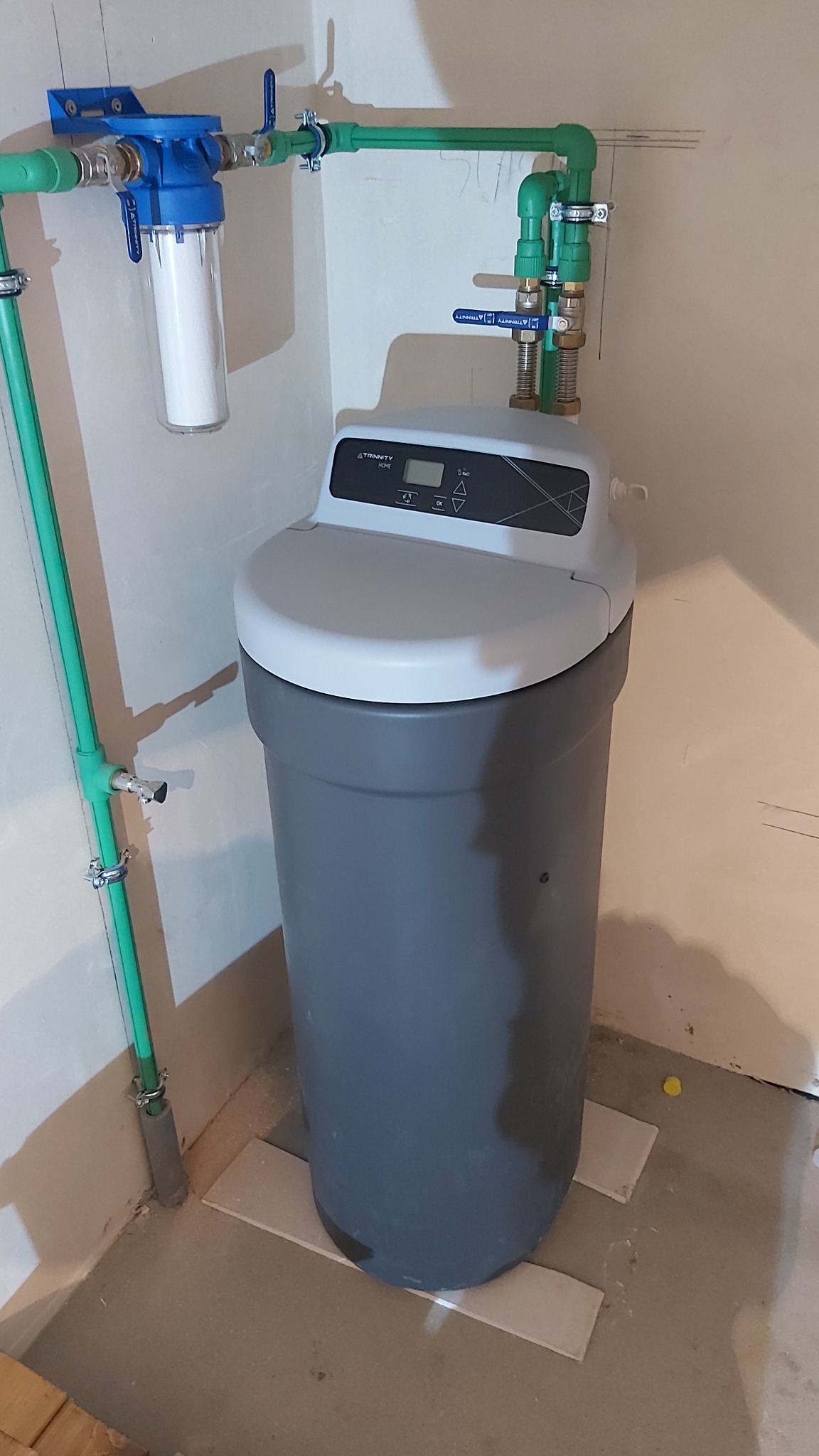 Zamontowany zmiękczacz wody Trinity Home z zielonymi rurami i filtrem wstępnym, zainstalowany w rogu pomieszczenia. Widoczne zawory i złączki.