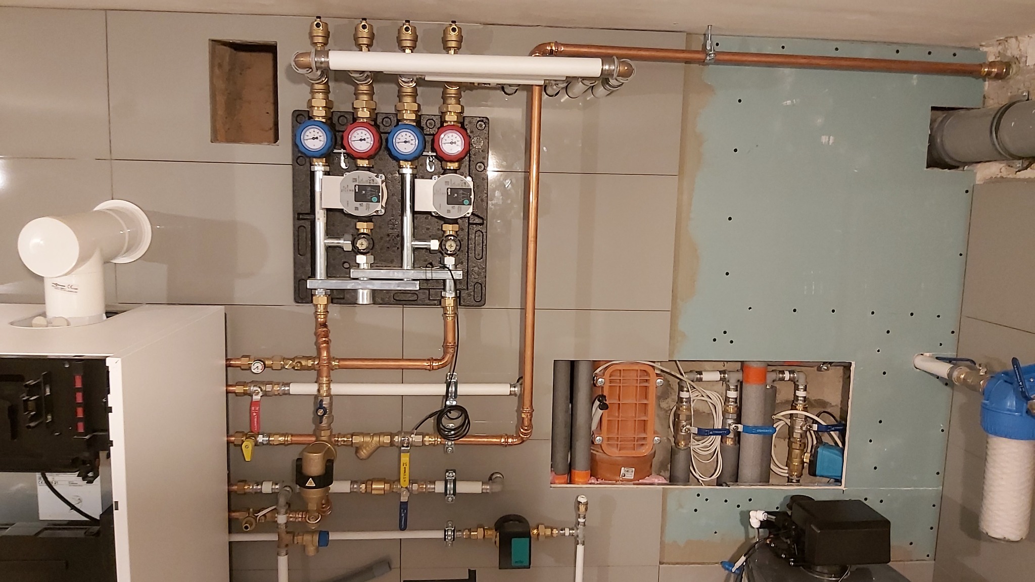 Kompleksowa instalacja grzewcza z piecem i rozdzielaczem, widoczne rury miedziane, manometry i pompy obiegowe na tle szarej ściany z płyt gipsowych i płytek.
