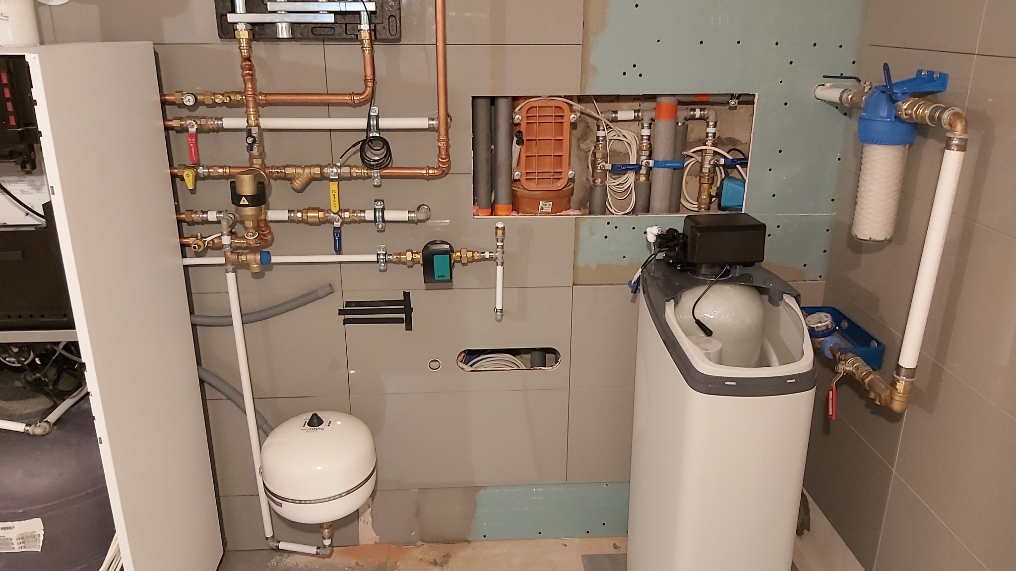 Złożona instalacja wodna z filtrem, rurami miedzianymi i białymi, zbiornikiem wyrównawczym oraz zmiękczaczem wody w pomieszczeniu wykończonym szarą płytką.